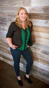Laura Michael en chemise verte et blazer noir, adossée à un mur en bois.